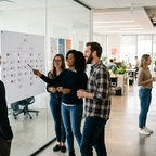 Mitarbeitende besprechen ein Organigramm im Büro, im Hintergrund unterhalten sich zwei Kollegen informell an der Kaffeemaschine
